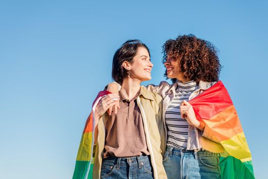 gay couple of lovely women embracing and looking into each other's eyes wrapped in the rainbow flag, symbol of the struggle for homosexual rights, concept of sexual freedom and racial diversity