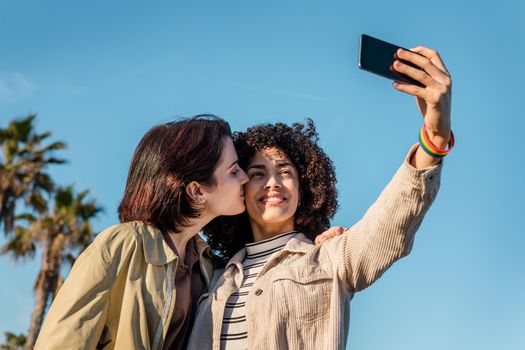 interracial young couple of homosexual women doing a selfie photo while kissing in a blue sky background with a mobile phone, concept of female friendship and racial diversity