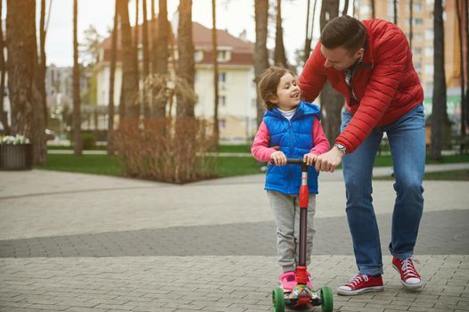 Handsome Caucasian man, loving and caring father walking beside his daughter riding a kick scooter while walking together in the city park. Happy family relationships and carefree childhood concept.