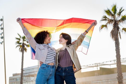 young lesbian couple of women embrace smiling under the rainbow flag, symbol of the struggle for gay rights, concept of sexual freedom and racial diversity