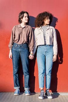 interracial gay couple of young women holding hands in front of a red wall, concept of sexual freedom and racial diversity, vertical photo