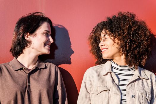 portrait of a young multiracial gay female couple looking into each other eyes holding hands in front of a red wall, concept of sexual freedom and racial diversity