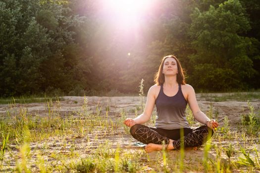 a woman practices yoga and meditates in the lotus position on the sand against the backdrop of trees.