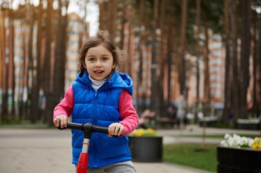 Waist length portrait of a Caucasian cute little girl riding a kick scooter in the city park on a sunny day in early spring. Healthy and active lifestyle, leisure activity, happy childhood concept