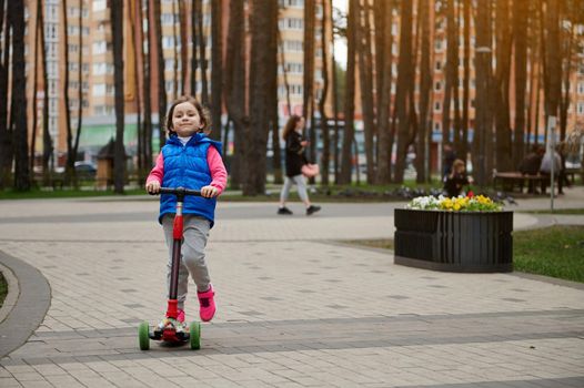 Full length portrait of a Caucasian cute little girl riding a kick scooter in the city park on a sunny day in early autumn or spring. Healthy and active lifestyle, leisure activity