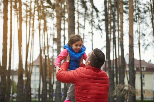 Loving caring father spends time with his daughter in a park, throws her up, enjoying her happy baby smile. Happy and carefree childhood, parenthood, Fathers Day concept, active and healthy lifestyle