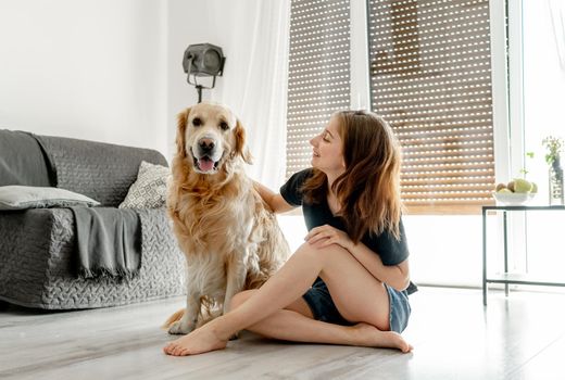 Pretty girl with golden retriever dog sitting on floor at home and smiling. Beautiful female teenager person with doggy pet in light room