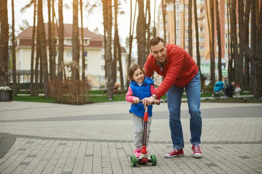 Happy friendly family of a charming loving father and his 5 year old daughter riding a push scooter outdoors in the forest park. Active lifestyle, parenthood, childhood, Father's Day concept