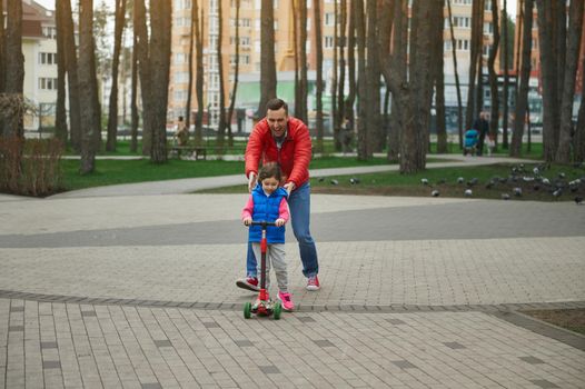 Cheerful dad running beside his cute daughter riding a push scooter, enjoying day off in a city park. Father's Day, happy childhood and fatherhood concept