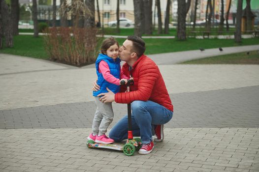 Happy loving father hugging his adorable little girl- beloved daughter and kissing her cheek while walking together in the city park, enjoying riding a push scooter on a beautiful early spring day