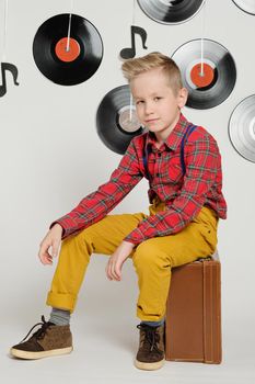 Retro disco 60s, 70s, 80s concept, funny boy wearing checked shirt, yellow trousers and stylish haircut sitting on a suitcase, background with music plate