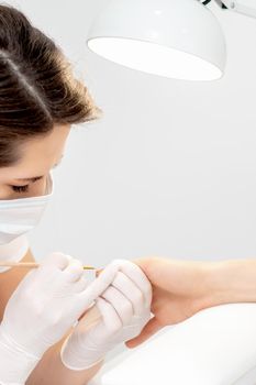 Manicure master with protective mask in rubber gloves applying beige nail polish on female nails in beauty salon