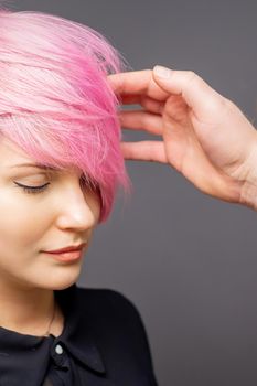 Hairdresser checking short pink hairstyle of young woman on gray background