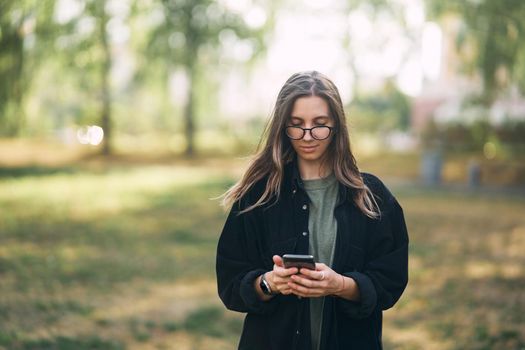 Young woman with glasses reading a message on her phone while standing in the park. High quality photo