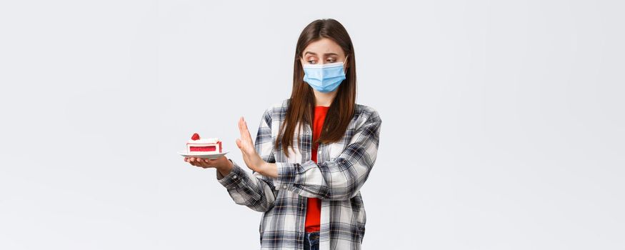 Coronavirus outbreak, lifestyle during social distancing and holidays celebration concept. Displeased and reluctant cute girl in medical mask refuse eat cake, show stop sign at dessert.