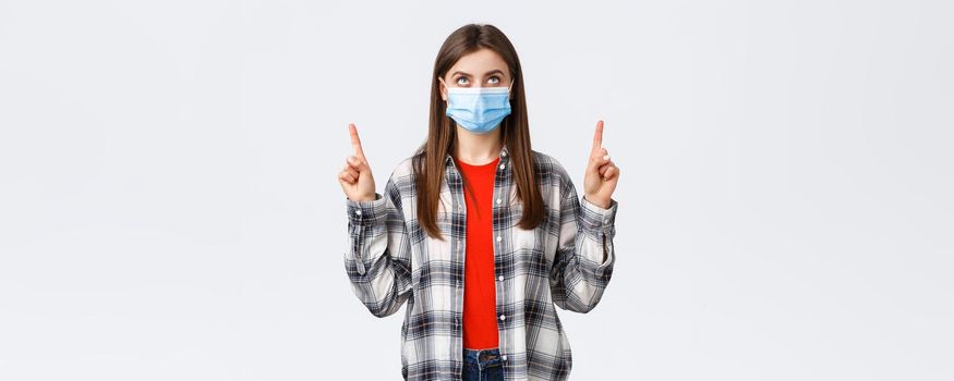 Coronavirus outbreak, leisure on quarantine, social distancing and emotions concept. Interested serious young woman in medical mask and casual outfit, looking and pointing up banner.