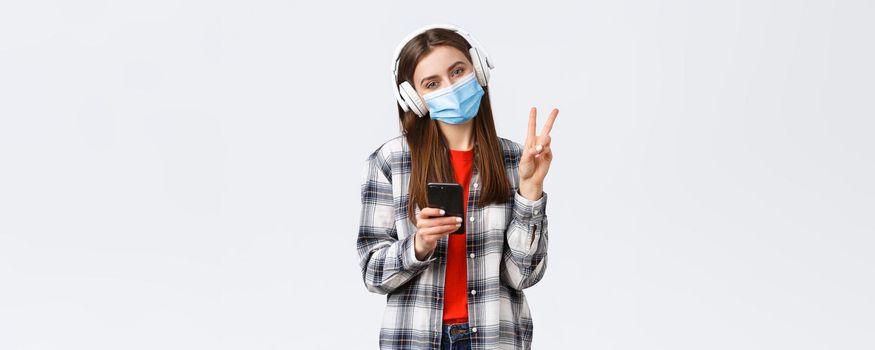 Social distancing, leisure and lifestyle on covid-19 outbreak, coronavirus concept. Cute silly teenage girl in medical mask and headphones, show peace sign, relaxing with music, hold mobile phone.