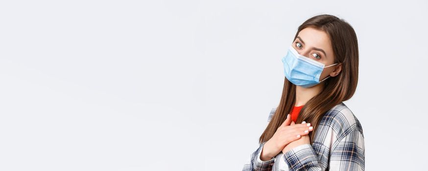 Coronavirus outbreak, leisure on quarantine, social distancing and emotions concept. Tender and touched young woman in medical mask, hold hands on heart feel grateful, white background.