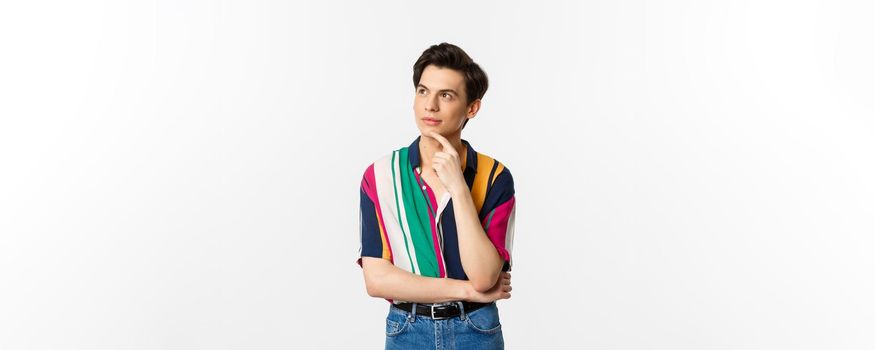 Portrait of thoughtful queer man in stylish shirt, looking at upper left corner and thinking, making choice, standing over white background.