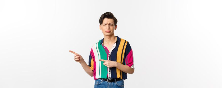Skeptical gay man looking with disdain and pointing fingers left, disapprove and dislike, standing over white background.