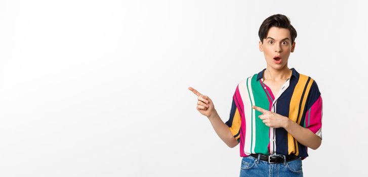 Amazed young queer man pointing fingers left, staring at camera with disbelief, asking question about product, standing over white background.