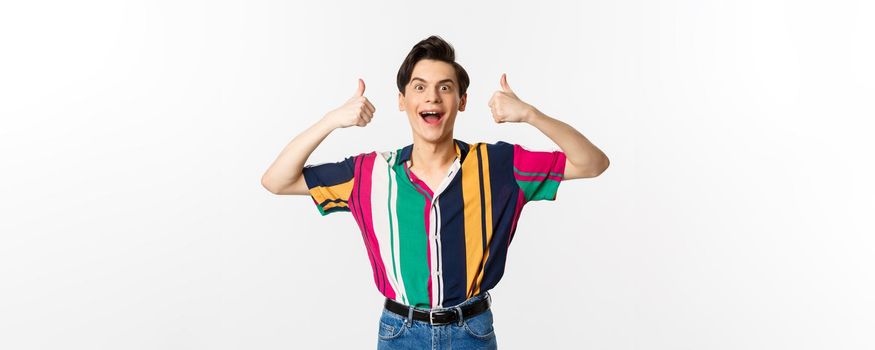 Excited and amazed young man showing thumbs up with impressed expression, standing over white background.