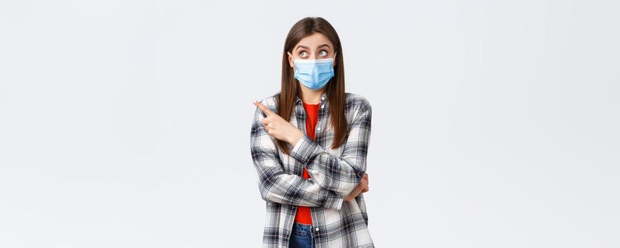 Coronavirus outbreak, leisure on quarantine, social distancing and emotions concept. Intrigued and surprised attractive girl in medical mask, casual clothes, pointing upper left corner interested.