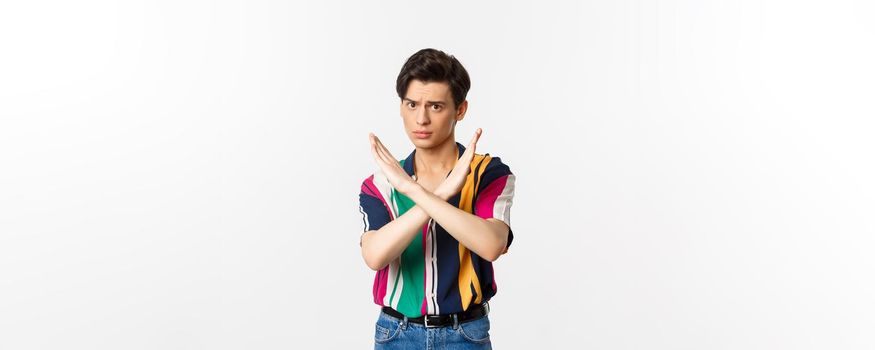 Disappointed young queer man telling no, showing stop gesture and forbid something bad, standing over white background.