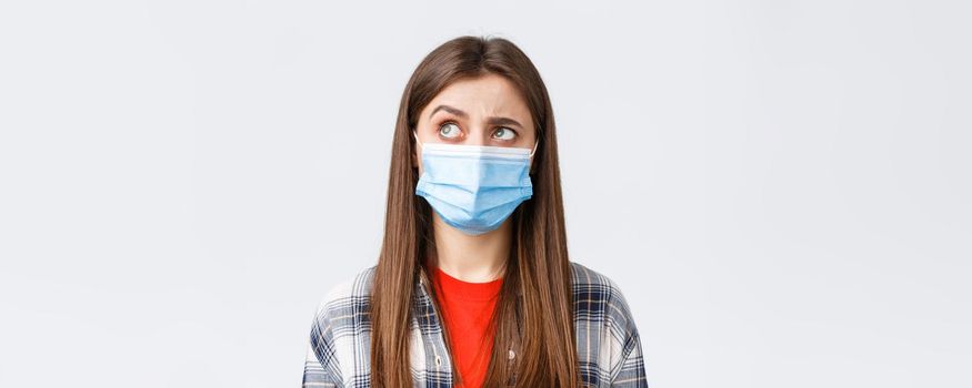 Coronavirus outbreak, leisure on quarantine, social distancing and emotions concept. Intrigued and hesitant young woman in medical mask, looking upper left corner reluctant, white background.