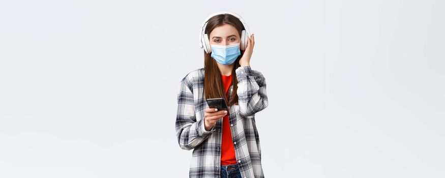 Social distancing, leisure and lifestyle on covid-19 outbreak, coronavirus concept. Cheerful smiling woman in medical mask, picking song from playlist smartphone, listen music in wireless headphones.