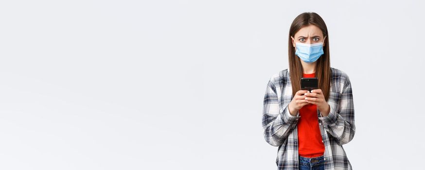 Different emotions, covid-19, social distancing and technology concept. Frustated and confused young woman in medical mask react to strange message, hold mobile phone, frowning camera.