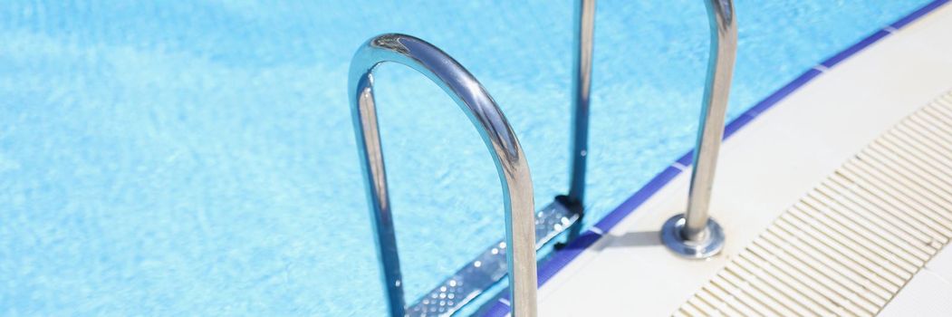 Close-up of ladder stainless handrails for descent into swimming pool safely. Clean blue water in hotel pool. Resort, summertime, sun, luxury hotel concept