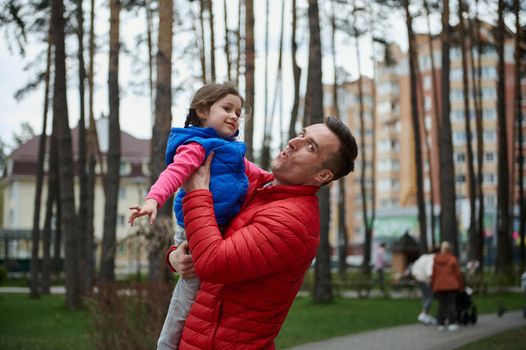 Handsome cheerful middle-aged Caucasian man - loving dad tosses his daughter - a little cute girl high up in a city park. Family and father's day, love, parents, children, carefree childhood concept