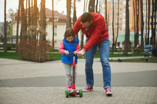 Caring father teaching his baby girl daughter riding a kick scooter in a city park. Active lifestyle, happy childhood and fatherhood, family relationships concept, leisure activity