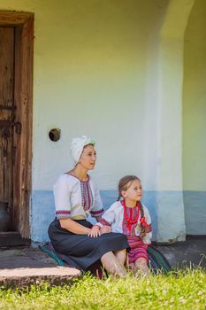 mother and daughter in Ukrainian national costumes are sitting near an old house