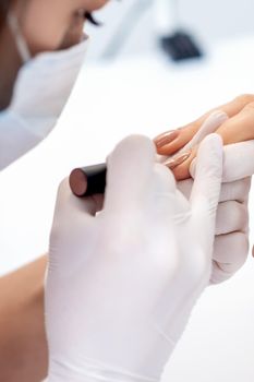 Manicure master in protective gloves applying beige nail polish on female nails in beauty salon