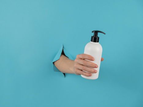 A woman's hand sticks out of a hole in a blue paper background and holds a skin lotion pump