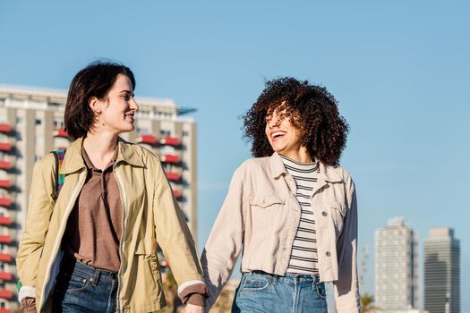 young interracial couple of women chatting and laughing while walking around the city holding hands, concept of sexual freedom and racial diversity