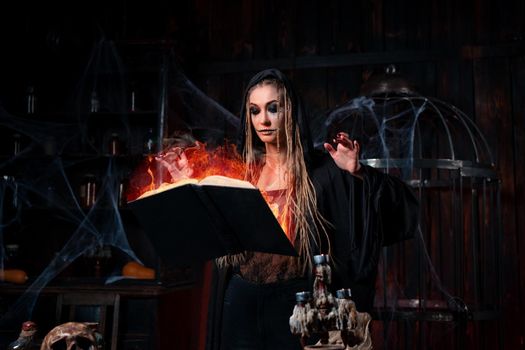 Halloween concept. Witch dressed black hood standing dark dungeon room use magic book fire effect for conjuring magic spell. Female necromancer wizard gothic interior with skull, cage, spider web