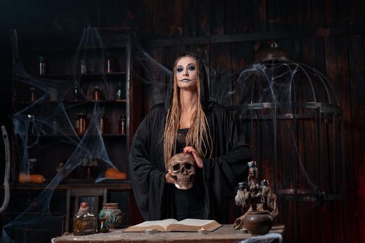 Halloween concept. Witch dressed black hood holding skull in hand standing dark dungeon room use magic book for conjuring magic spell. Female necromancer wizard gothic interior