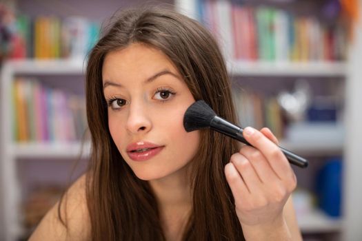 A teenager looks straight into the camera and powders her cheeks with a makeup brush. The art of painting your face yourself.