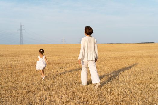 Little girl with her mother dressed white clothes walking wheat field back view blue sky background. Daughter with mom run nature countryside view Freedom or happy family concept