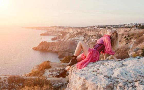 Side view a Young beautiful sensual woman in a red long dress posing on a volcanic rock high above the sea during sunset. Girl on the nature on blue sky background. Fashion photo