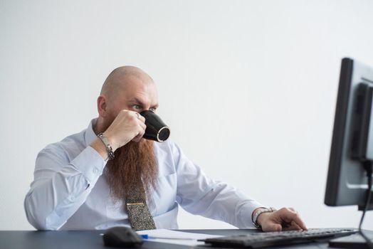 Focused bald man with a red beard drinks coffee and works at the computer. Male manager in a white shirt at a personal computer in the office