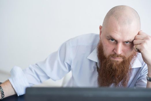 A focused bald man with a red beard stares intently at the camera. Male manager in a white shirt is angry at work