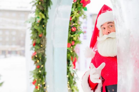 Santa claus peeks out of Christmas decorations outdoors.