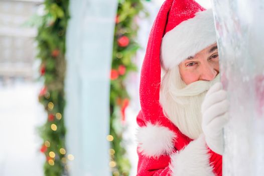 Santa claus peeks out of Christmas decorations outdoors.