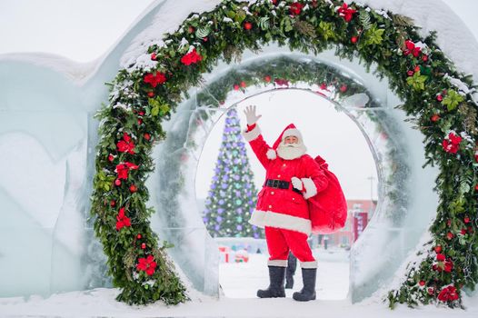 Santa Claus posing with a bag of gifts on the background of Christmas decorations outdoors.