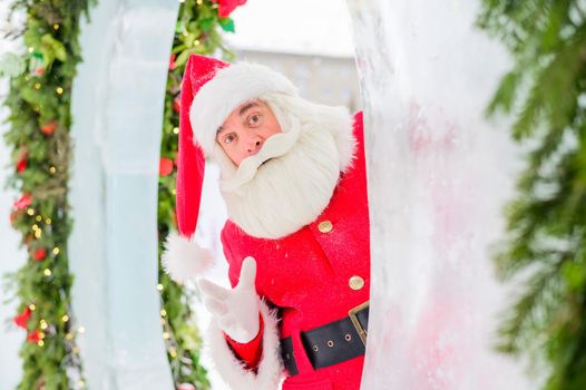 Santa claus peeks out of Christmas decorations outdoors.