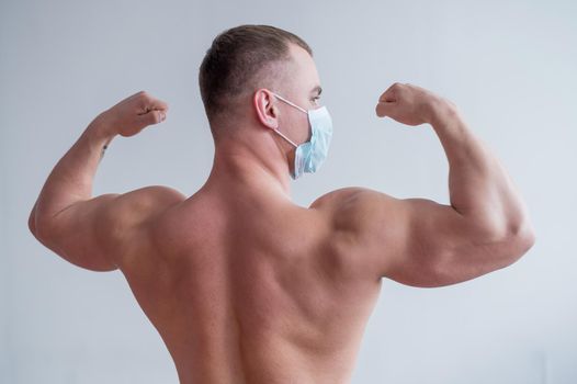 Male bodybuilder in a medical mask stands with his back. The guy goes in for sports in quarantine. Respiratory protection. Shows biceps